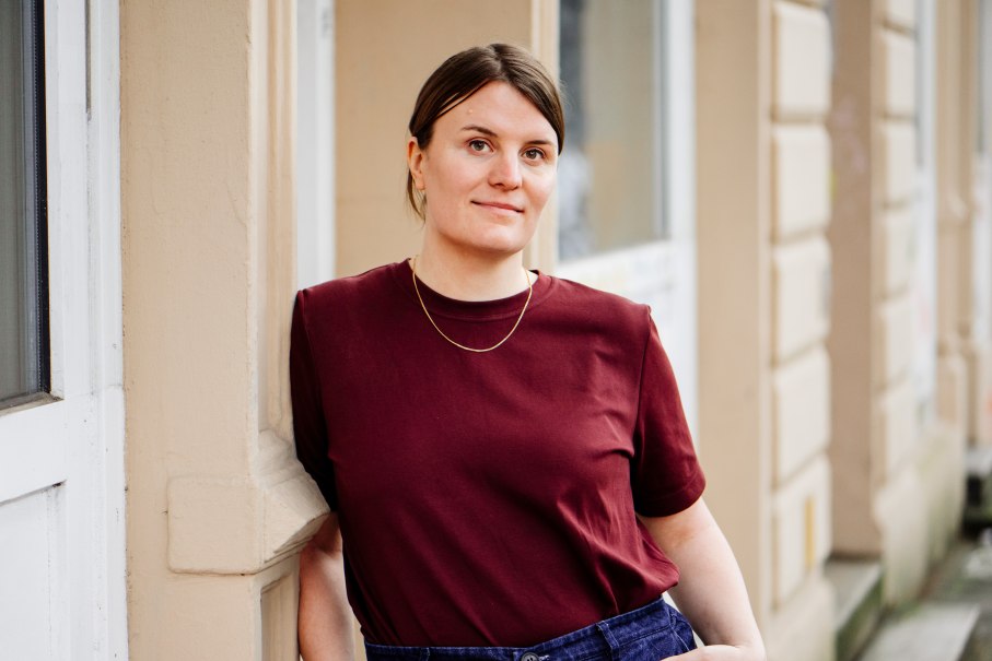 Eine Frau in einem weinroten T-Shirt lehnt entspannt an einer beigen Wand. Sie tr&auml;gt eine schlichte Halskette und blickt in die Kamera., &copy; Asja Caspari