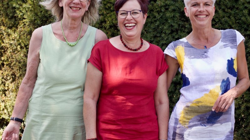Drei Frauen in bunten Sommerkleidern stehen lächelnd vor einer grünen Hecke., © Ursel Krieg Drei Frauen in bunten Sommerkleidern stehen lächelnd vor einer grünen Hecke., © Ursel Krieg