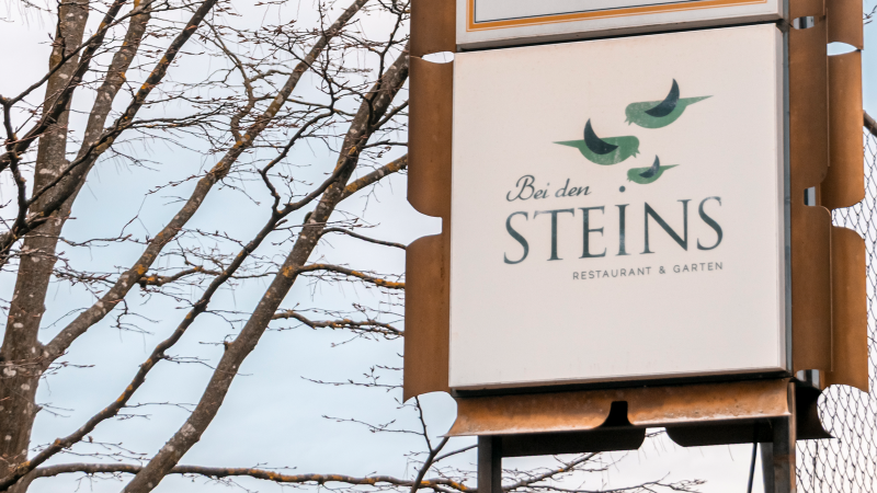 A sign for the restaurant 'Bei den Steins' with birds as a logo, in front of a bare tree and blue sky., &copy; Stuttgart-Marketing GmbH, Sarah Schmid
