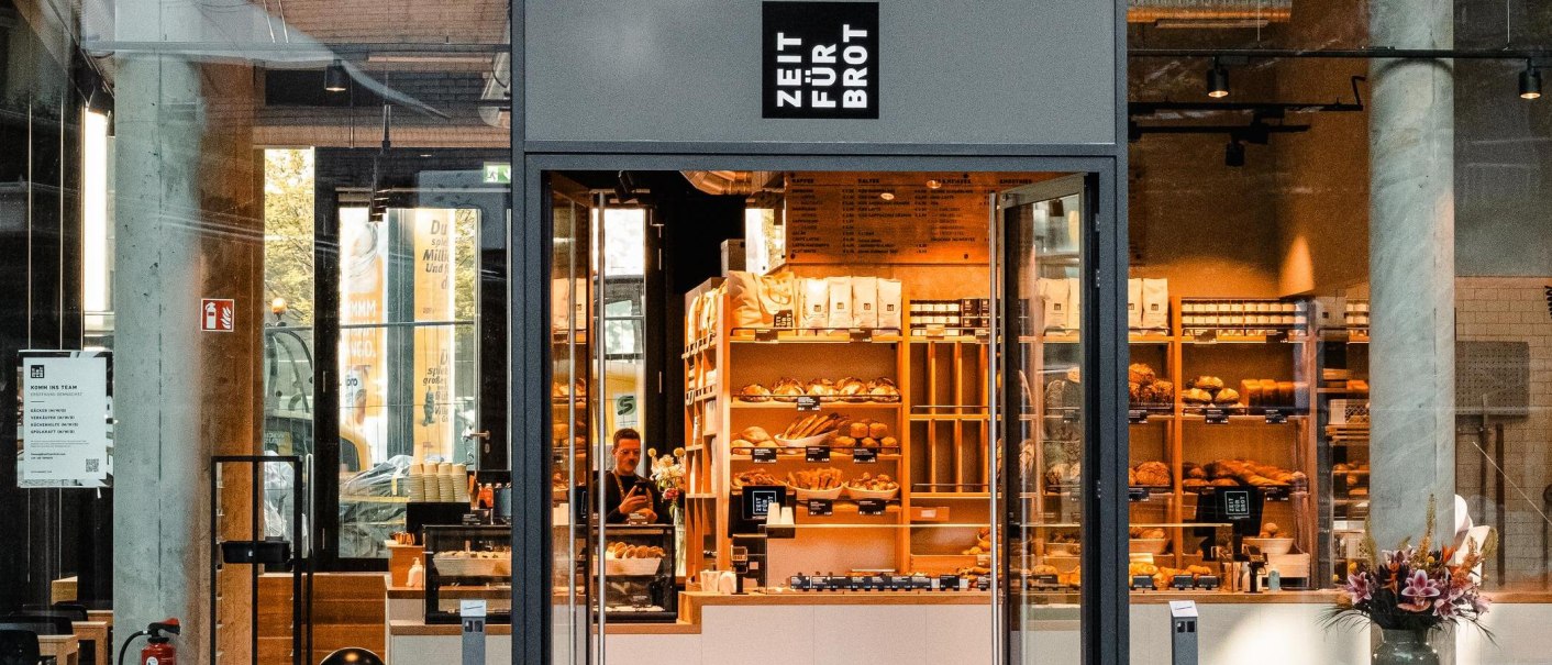 Eingang einer Bäckerei mit dem Schild 'Zeit für Brot'. Innen sind Regale mit Brot und Gebäck zu sehen., © Zeit für Brot Stuttgart