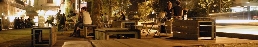 Nachtaufnahme der Theodor-Heuss-Stra&szlig;e in Stuttgart. Menschen sitzen auf Holzb&auml;nken unter B&auml;umen, umgeben von beleuchteten Geb&auml;uden und Stra&szlig;enlaternen., &copy; Stuttgart-Marketing GmbH, Marc Gilardone