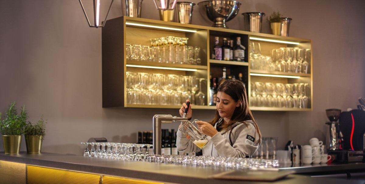 Frau zapft Bier an einer modernen Bar mit beleuchteten Regalen und Gläsern. Elegante Beleuchtung und Dekoration., © Novum Hospitality