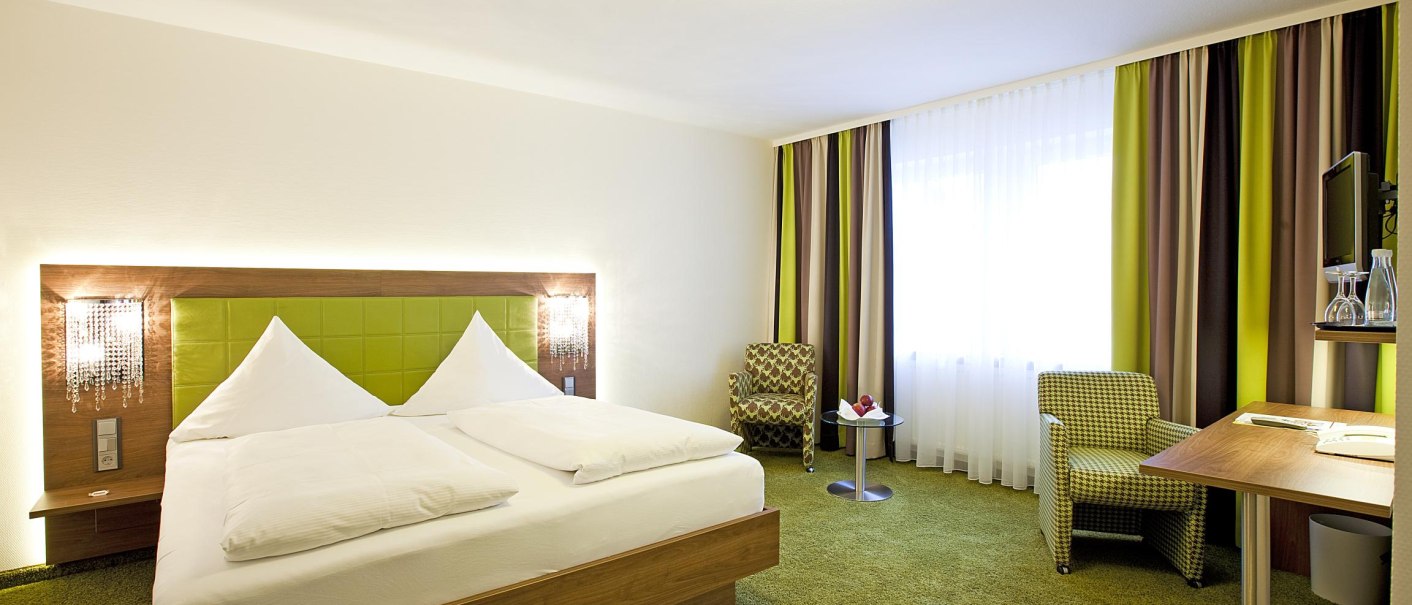 Modernes Hotelzimmer mit grünem Teppich, Doppelbett, Schreibtisch, zwei Sesseln und Fenster mit Vorhängen., © Hotel Hirsch