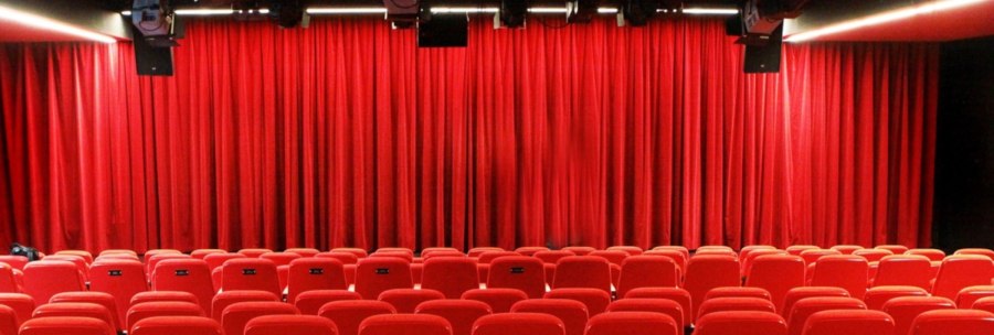 Empty theater hall with red chairs and closed red curtain, lighting and loudspeakers on the ceiling., © Sabine Haymann Empty theater hall with red chairs and closed red curtain, lighting and loudspeakers on the ceiling., © Sabine Haymann