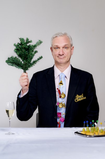Mann in Anzug mit Weihnachtsdeko hält Tannenzweig. Vor ihm Sektglas und Snacktablett. Hintergrund neutral., © Rosenau Kultur e.V.