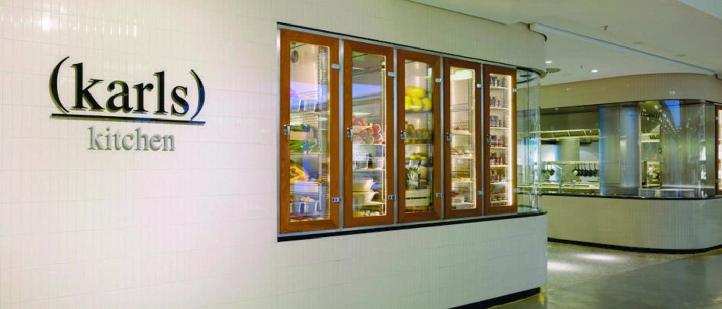 Entrance to "karls kitchen" with illuminated glass cabinets full of food and a view of the modern kitchen., © karls kitchen, Stuttgart