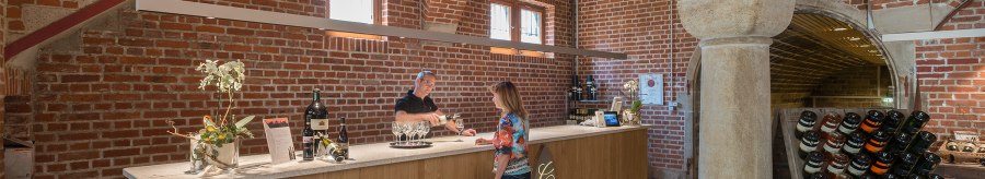 A bar in a wine cellar with brick walls. A man pours wine for a woman. Wine bottles are displayed on the counter., © Collegium Wirtemberg