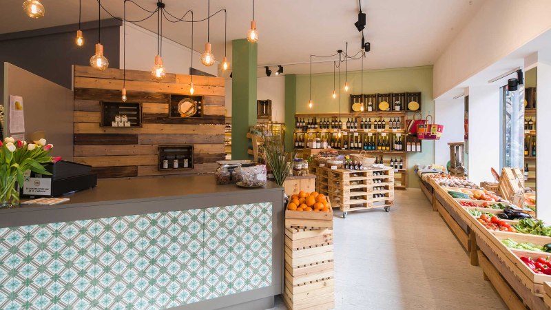 Moderner Feinkostladen mit Holzregalen, frischem Obst und Gemüse. Hängelampen und eine Theke mit dekorativen Fliesen verleihen eine gemütliche Atmosphäre., © Benjamin Stollenberg