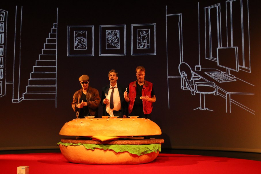 Drei Männer stehen hinter einem riesigen Hamburger-Requisit auf einer Bühne. Der Hintergrund zeigt gezeichnete Möbel und Bilderrahmen., © Sabine Haymann Drei Männer stehen hinter einem riesigen Hamburger-Requisit auf einer Bühne. Der Hintergrund zeigt gezeichnete Möbel und Bilderrahmen., © Sabine Haymann