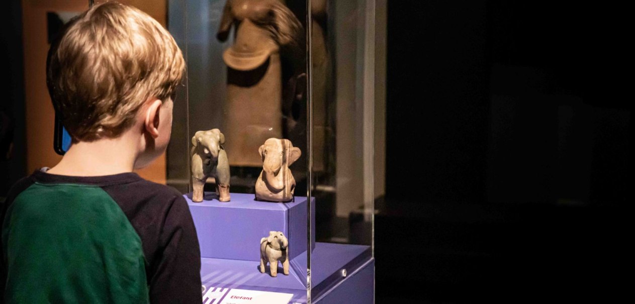 Ein Junge betrachtet aufmerksam Elefantenfiguren in einer Museumsvitrine. Die Ausstellung scheint Teil eines Ferienprogramms zu sein., &copy; Linden-Museum Stuttgart