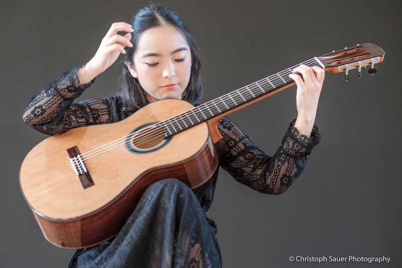 Eine Frau in schwarzer Spitzenkleidung spielt konzentriert auf einer klassischen Gitarre vor einem neutralen Hintergrund., &copy; Staatliche Hochschule f&uuml;r Musik und Darstellende Kunst