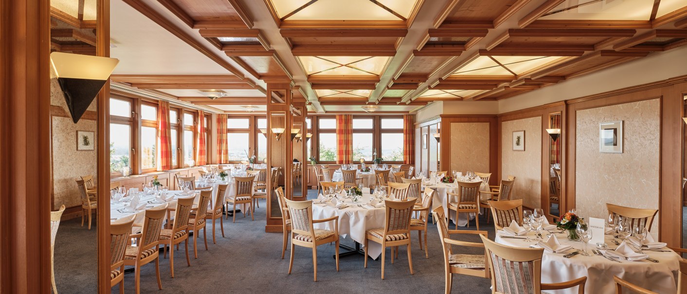 An elegant dining room with set tables, wood paneling, and large windows. The tables are adorned with white tablecloths and floral arrangements., © Badhotel Restaurant Stauferland An elegant dining room with set tables, wood paneling, and large windows. The tables are adorned with white tablecloths and floral arrangements., © Badhotel Restaurant Stauferland