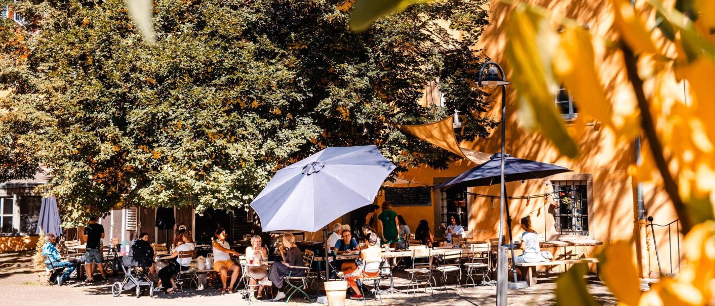 Menschen genießen das sonnige Wetter in einem Café im Freien, umgeben von Bäumen und Sonnenschirmen. Eine entspannte Atmosphäre herrscht vor., © Stuttgart-Marketing GmbH, Sarah Schmid Menschen genießen das sonnige Wetter in einem Café im Freien, umgeben von Bäumen und Sonnenschirmen. Eine entspannte Atmosphäre herrscht vor., © Stuttgart-Marketing GmbH, Sarah Schmid