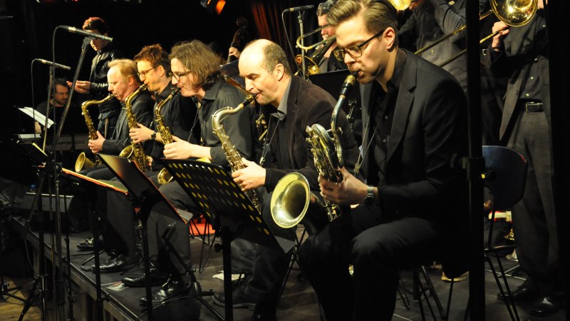 Eine Big Band auf der Bühne, Musiker spielen Saxophone und Posaunen. Die Bühne ist in warmes Licht getaucht, Notenständer sind sichtbar., © Pressefoto