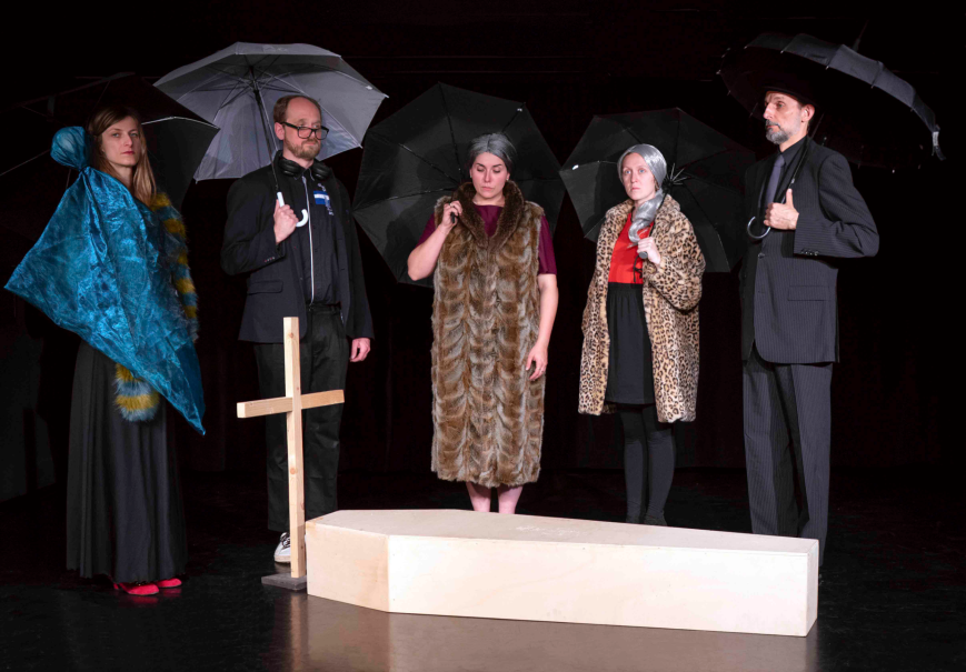 Fünf Personen in schwarzer Kleidung stehen mit Regenschirmen um einen Sarg. Ein Holzkreuz ist sichtbar., © Gerhard Killet Fünf Personen in schwarzer Kleidung stehen mit Regenschirmen um einen Sarg. Ein Holzkreuz ist sichtbar., © Gerhard Killet