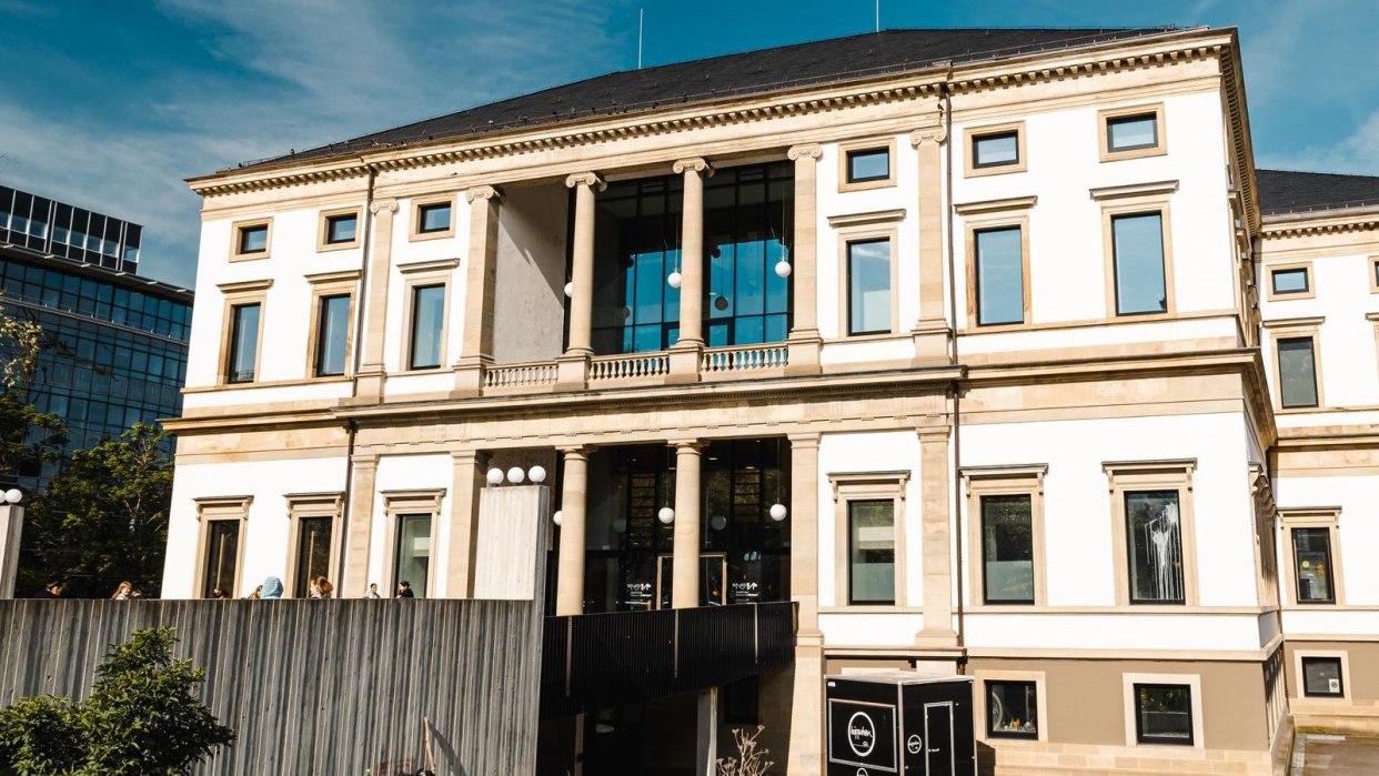 Ein historisches Gebäude mit Säulen und großen Fenstern, umgeben von moderner Architektur und Bäumen, unter einem klaren blauen Himmel., © Stuttgart Marketing GmbH, Sarah Schmid Ein historisches Gebäude mit Säulen und großen Fenstern, umgeben von moderner Architektur und Bäumen, unter einem klaren blauen Himmel., © Stuttgart Marketing GmbH, Sarah Schmid