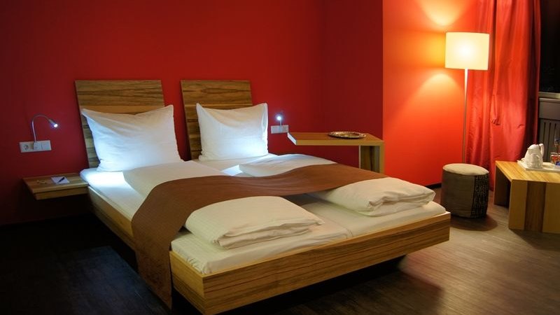 Modernes Hotelzimmer mit roter Wand, Doppelbett, Nachttischlampen und Stehlampe. Elegante Einrichtung mit Holzmöbeln und warmem Licht., © AMH Airport-Messe-Hotel Modernes Hotelzimmer mit roter Wand, Doppelbett, Nachttischlampen und Stehlampe. Elegante Einrichtung mit Holzmöbeln und warmem Licht., © AMH Airport-Messe-Hotel