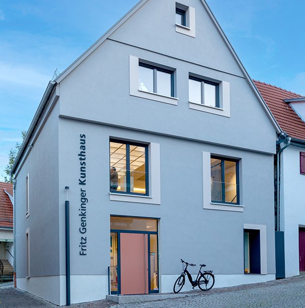 The Fritz Genkinger Kunsthaus has a gray facade with large windows. A bicycle is parked in front of it on the paved path., © Freundeskreis Fritz Genkinger e.V. The Fritz Genkinger Kunsthaus has a gray facade with large windows. A bicycle is parked in front of it on the paved path., © Freundeskreis Fritz Genkinger e.V.