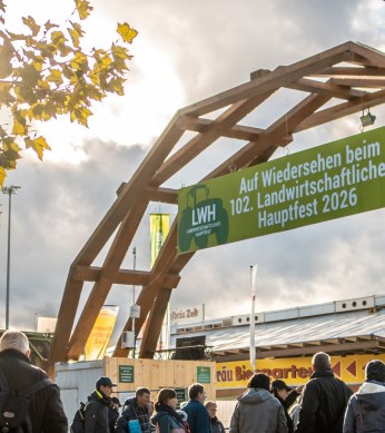 Besucher betreten das Landwirtschaftliche Hauptfest 2026 unter einem Holzbogen mit Banner. Herbstliche Atmosph&auml;re mit gelben Bl&auml;ttern und Wolken., &copy; Daniel Schneider, 2022