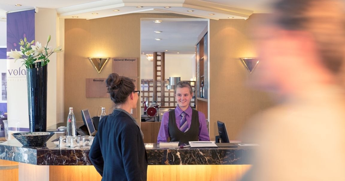 A hotel reception desk where a staff member is talking to a guest; elegant decor featuring flowers and a marble counter; a blurred figure in the foreground., &copy; Mercure Hotel Stuttgart Gerlingen