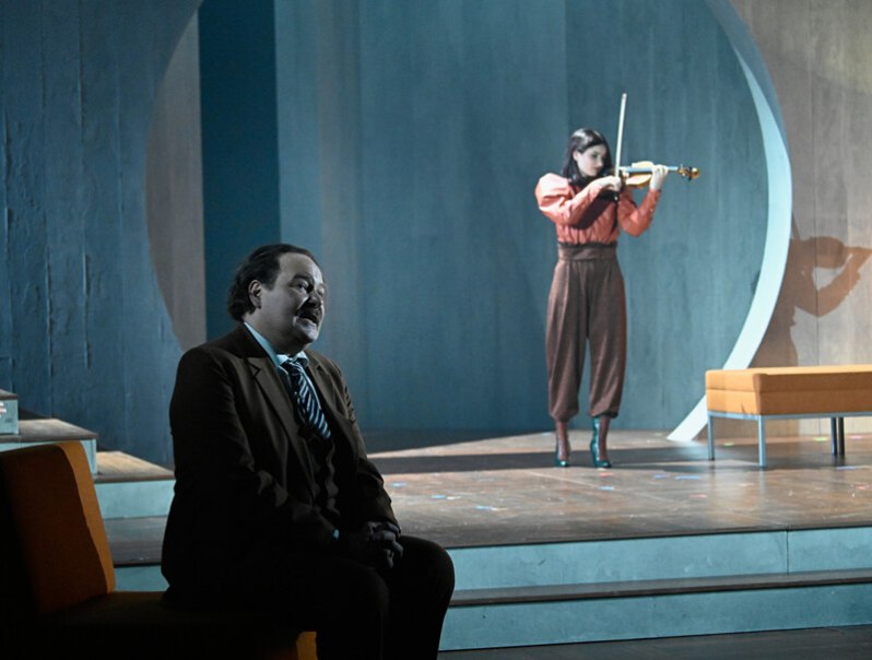 Ein Mann im Anzug sitzt auf einer B&uuml;hne, w&auml;hrend eine Frau im Hintergrund Geige spielt. Die Szene wirkt dramatisch und konzentriert., &copy; W&uuml;rttembergische Staatstheater Stuttgart