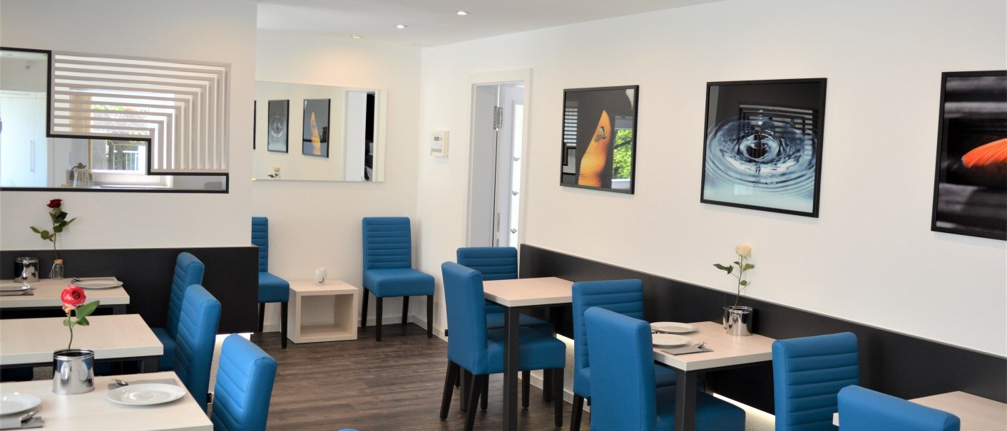 Moderner Frühstücksraum mit blauen Stühlen, weißen Tischen, Blumen und Kunstwerken an den Wänden. Helle Beleuchtung und Holzfußboden., © Hotel BaWü Moderner Frühstücksraum mit blauen Stühlen, weißen Tischen, Blumen und Kunstwerken an den Wänden. Helle Beleuchtung und Holzfußboden., © Hotel BaWü