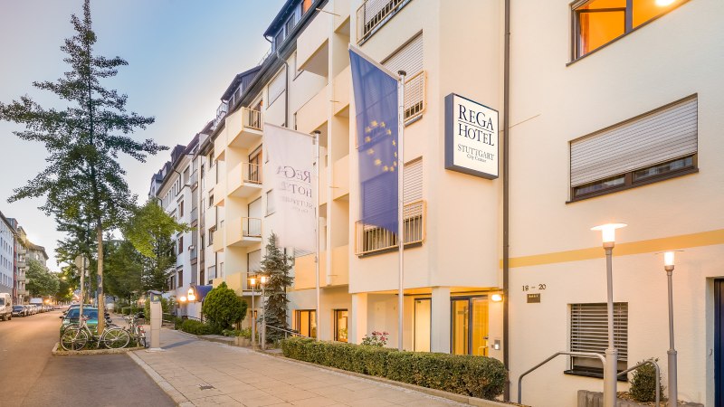 Exterior view of the Rega Hotel in Stuttgart. Multi-storey building with flags and illuminated street lamps. Quiet street with parked cars., © TOMAS