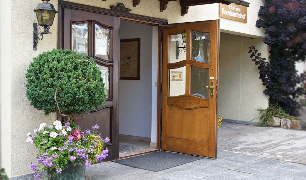 Open wooden door of a hotel with a potted plant and colorful flowers in front of it., © Hotel Reinhardtshof Garni Open wooden door of a hotel with a potted plant and colorful flowers in front of it., © Hotel Reinhardtshof Garni