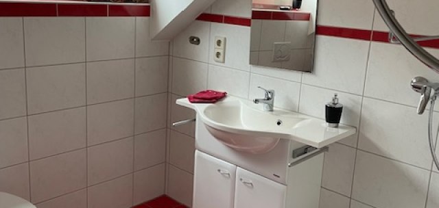 Badezimmer mit roten Bodenfliesen, weißem Waschbecken, Spiegel und Fenster mit Jalousie. Weiße Wandfliesen mit rotem Streifen., © Weinhof am Württemberg Badezimmer mit roten Bodenfliesen, weißem Waschbecken, Spiegel und Fenster mit Jalousie. Weiße Wandfliesen mit rotem Streifen., © Weinhof am Württemberg