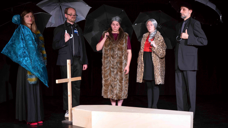Fünf Personen in schwarzer Kleidung stehen mit Regenschirmen um einen Sarg. Ein Holzkreuz ist sichtbar., © Gerhard Killet Fünf Personen in schwarzer Kleidung stehen mit Regenschirmen um einen Sarg. Ein Holzkreuz ist sichtbar., © Gerhard Killet