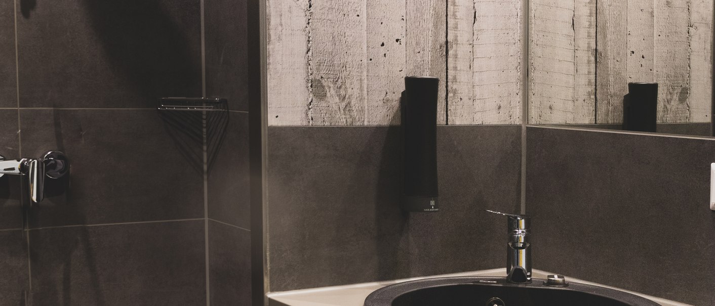 Modernes Badezimmer mit dunklen Fliesen, einem Waschbecken und einem Spiegel. An der Wand hängt ein Seifenspender., © Das Schlafwerk - DQuadrat Living GmbH