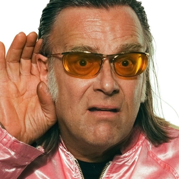 Person with orange-tinted glasses and a pink jacket holds his hand to his ear as if he wants to hear better., &copy; Theaterhaus Stuttgart e.V.