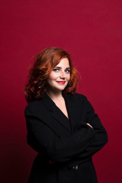 Woman with red hair and black blazer stands smiling in front of a red background., &copy; Rosenau Kultur e.V.