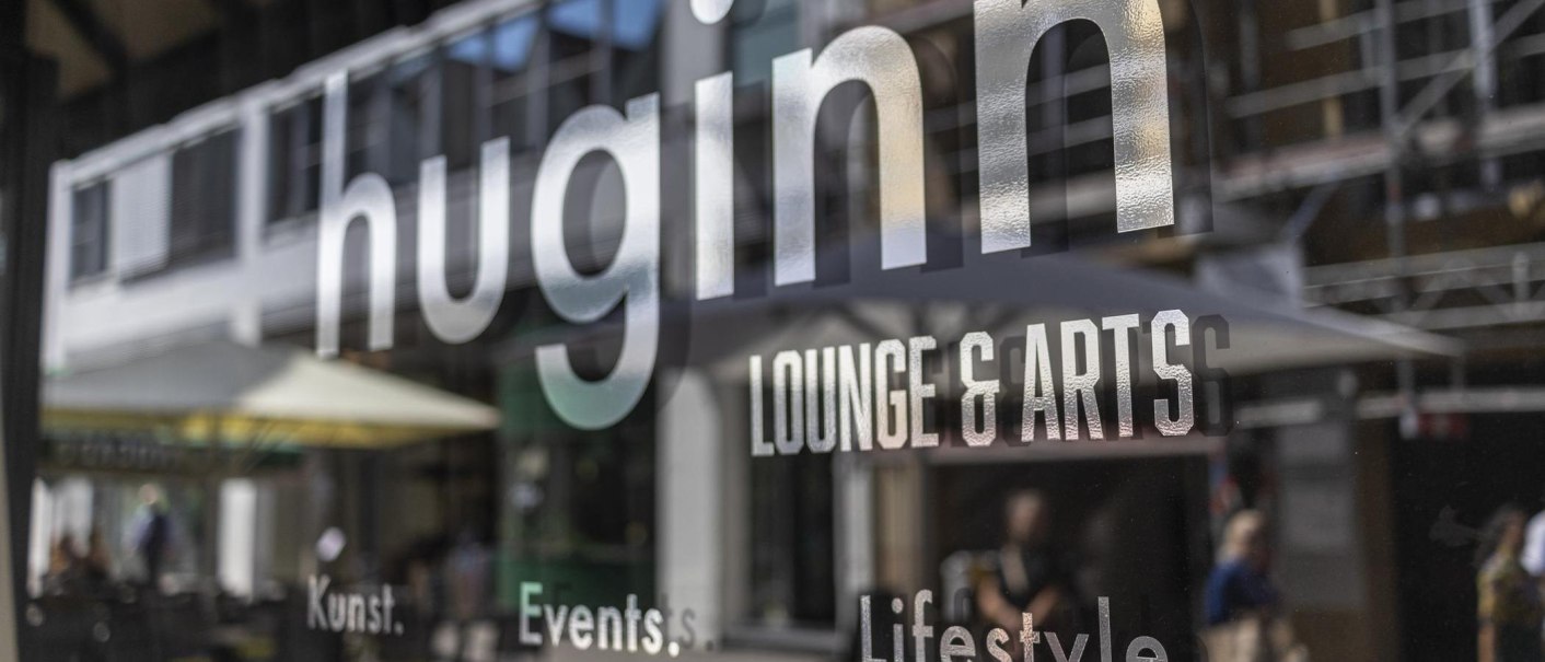 Spiegelung des Schriftzugs 'huginn LOUNGE & ARTS' auf einer Glasoberfläche, im Hintergrund unscharfe Straßenszene., © SMG, Sarah Schmid