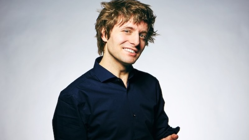 Ein Mann mit zerzaustem Haar lächelt in die Kamera, trägt ein dunkelblaues Hemd vor grauem Hintergrund., © Renitenztheater Stuttgart e.V.