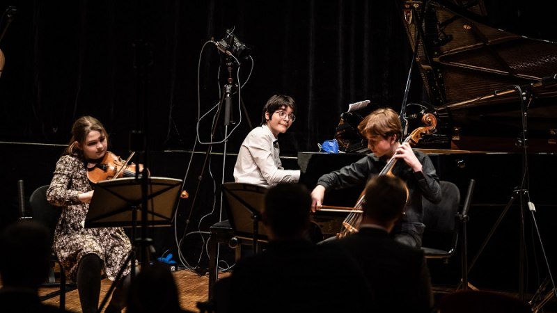Drei junge Musiker spielen Violine, Klavier und Cello auf einer Bühne vor Publikum. Im Hintergrund steht ein Flügel., © unbekannt Drei junge Musiker spielen Violine, Klavier und Cello auf einer Bühne vor Publikum. Im Hintergrund steht ein Flügel., © unbekannt