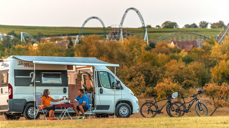 Wohnmobil mit Markise auf Wiese, zwei Personen am Tisch, Fahrräder daneben, Achterbahnen im Hintergrund, herbstliche Landschaft., © Erlebnispark Tripsdrill GmbH & Co. KG Wohnmobil mit Markise auf Wiese, zwei Personen am Tisch, Fahrräder daneben, Achterbahnen im Hintergrund, herbstliche Landschaft., © Erlebnispark Tripsdrill GmbH & Co. KG