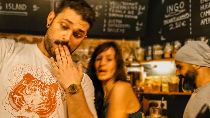 Drei Personen in einer Bar, eine mit Hand vor dem Mund, eine lächelnd, eine mit Mütze. Im Hintergrund eine Getränketafel., © Café Galao