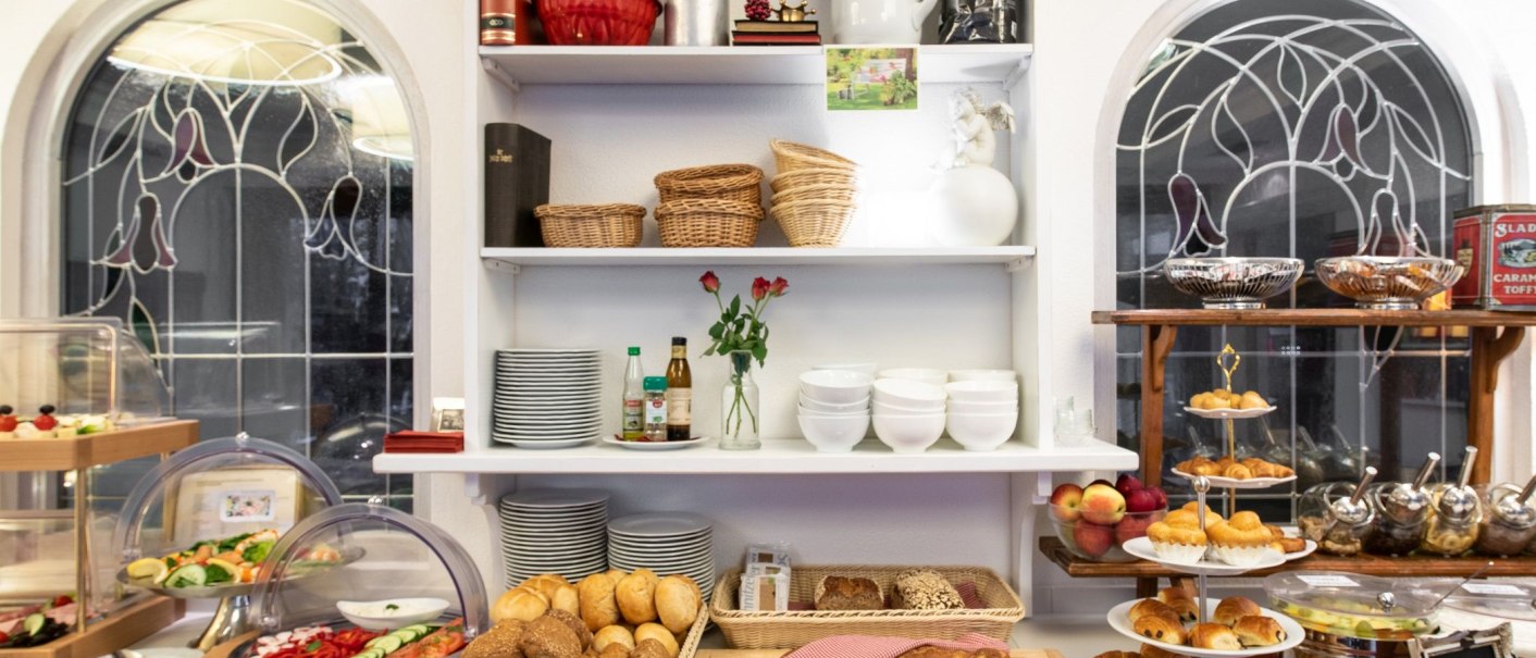 A rich breakfast buffet with bread, pastries, fruit and crockery. Decorative windows can be seen in the background., © Hotel Weinstadt