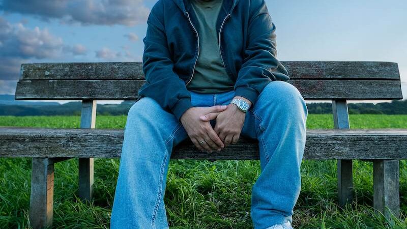 Ein Mann sitzt auf einer Holzbank in einer Wiese, trägt Jeans und eine Jacke. Der Himmel ist blau mit Wolken., © SBEntertainment GmbH & Co. KG Ein Mann sitzt auf einer Holzbank in einer Wiese, trägt Jeans und eine Jacke. Der Himmel ist blau mit Wolken., © SBEntertainment GmbH & Co. KG