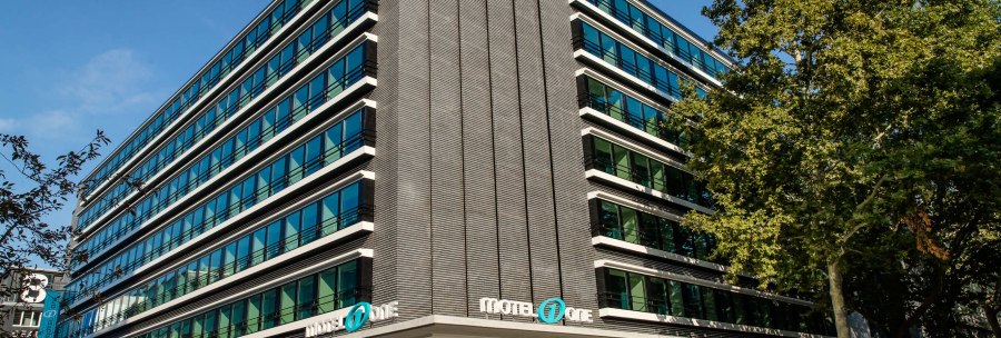 Modern building with glass facade and Motel One logo, surrounded by trees, under a blue sky., &copy; Motel One - Stuttgart Hauptbahnhof