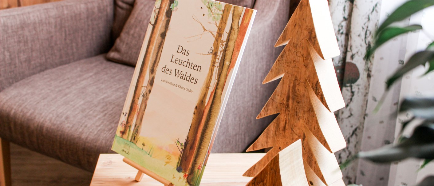 Ein Buch mit dem Titel 'Das Leuchten des Waldes' steht auf einem Tisch neben einer Holzdekoration in Form eines Tannenbaums., © TOMAS