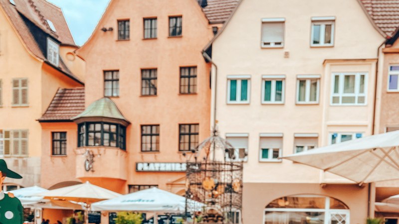 The Hans im Gl&uuml;ck fountain in front of historic buildings. Parasols and figures can be seen in the foreground. The houses have red roofs and many windows., &copy; SMG, takemyhearteverywhere