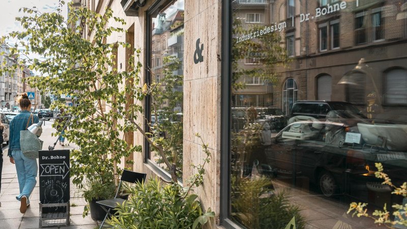 Zwei Personen gehen an einem Gesch&auml;ft mit der Aufschrift 'Dr. Bohne' vorbei. Die Stra&szlig;e ist von Autos ges&auml;umt, und Pflanzen stehen vor dem Schaufenster., &copy; SMG, Sarah Schmid