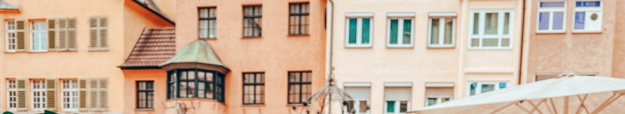 The Hans im Gl&uuml;ck fountain in front of historic buildings. Parasols and figures can be seen in the foreground. The houses have red roofs and many windows., &copy; SMG, takemyhearteverywhere