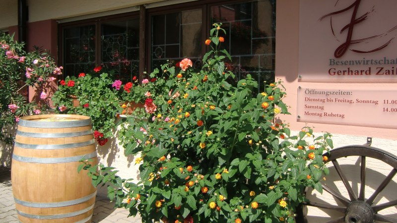 Eingang der Besenwirtschaft Gerhard Zaiß mit blühenden Pflanzen, einem Weinfass und einem Schild mit Öffnungszeiten., © Simone Mack