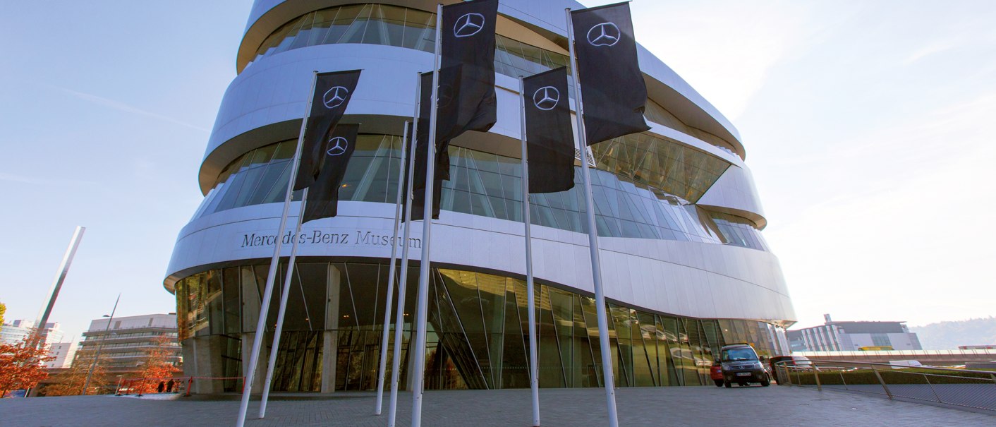 The Mercedes-Benz Museum with its modern architecture and several flags in the foreground. The sky is clear and blue., &copy; SMG, Achim Mende