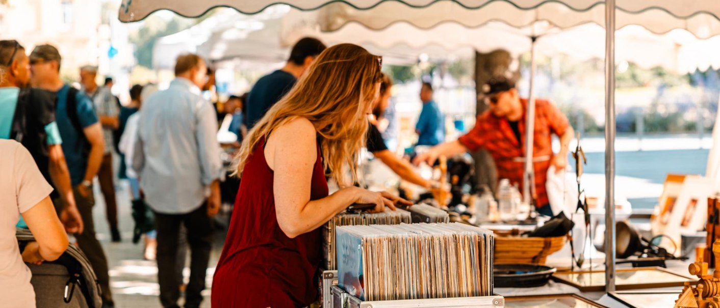 Eine Frau durchsucht Schallplatten auf einem belebten Flohmarkt unter Sonnenschirmen. Menschen im Hintergrund betrachten weitere Stände., © Stuttgart Marketing GmbH, Sarah Schmid