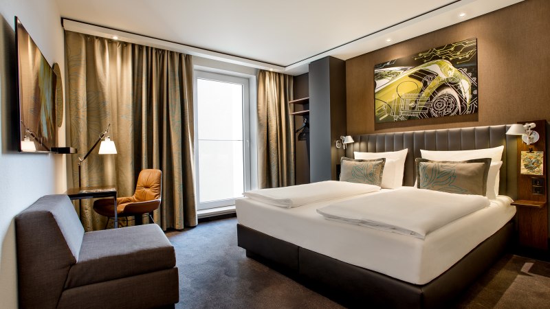 Modernes Hotelzimmer mit Doppelbett, Sofa, Schreibtisch und großem Fenster. An der Wand hängt ein Kunstwerk mit einem Auto-Motiv., © TOMAS