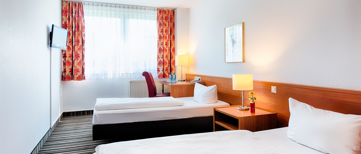 A bright twin room with two single beds, a nightstand with a lamp, a desk, a TV, and red curtains., &copy; ACHAT Hotels Deutschland